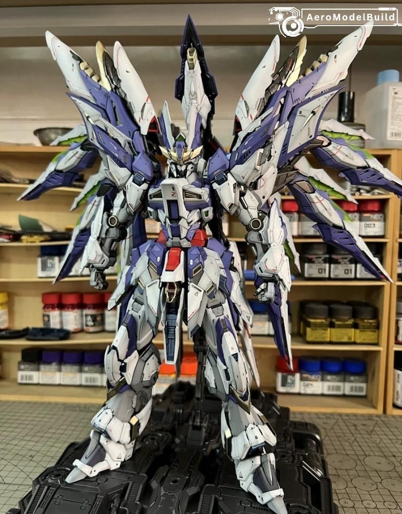 May include: A highly detailed model robot with large wings, predominantly white, blue, and grey, with red and gold highlights. The robot stands on a black base, displaying intricate mechanical details and a complex design, suitable for collectors.