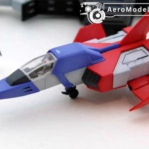 May include: AeroModelBuild model airplane in red, blue, and white. The toy plane has a cockpit and wings, and is next to a small figurine. The plane is on a white surface.