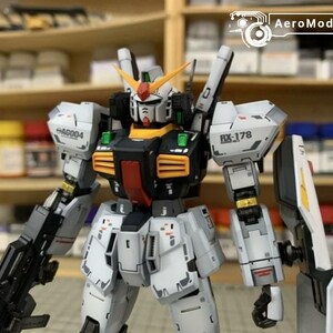 Pode incluir: Um modelo detalhado de um rob&ocirc; branco, preto e amarelo com uma arma. O rob&ocirc; tem o texto "AG004" e "RX-178" impresso. O rob&ocirc; est&aacute; em frente a uma prateleira com v&aacute;rias garrafas.