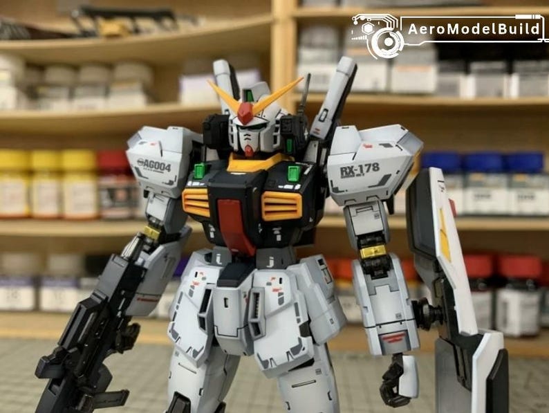 Pode incluir: Um modelo detalhado de um rob&ocirc; branco, preto e amarelo com uma arma. O rob&ocirc; tem o texto "AG004" e "RX-178" impresso. O rob&ocirc; est&aacute; em frente a uma prateleira com v&aacute;rias garrafas.