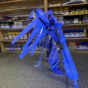 May include: A transparent blue model robot, featuring large wing-like elements. The model is positioned on a cutting mat, with paint pots and tools visible on shelves in the background. The AeroModelBuild logo is present.