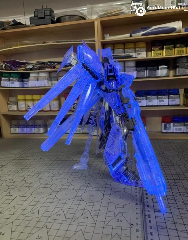 May include: A transparent blue model robot, featuring large wing-like elements. The model is positioned on a cutting mat, with paint pots and tools visible on shelves in the background. The AeroModelBuild logo is present.