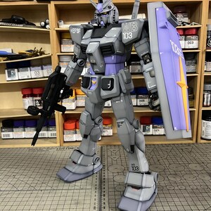 May include: A detailed model of a Gundam robot, primarily gray with purple and white accents. The robot is holding a black rifle and a shield with the text "103". The model is standing on a cutting mat with measurement markings.
