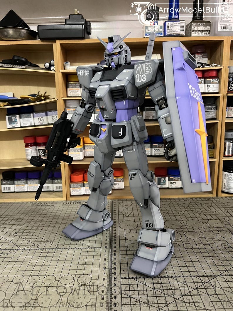 May include: A detailed model of a Gundam robot, primarily gray with purple and white accents. The robot is holding a black rifle and a shield with the text "103". The model is standing on a cutting mat with measurement markings.
