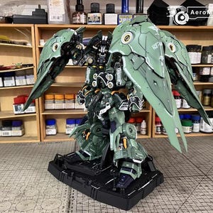 May include: A detailed, green and black model robot with large wing-like structures. The model is on a black base, showcasing intricate mechanical details. The background includes shelves with various paints and model-building supplies.