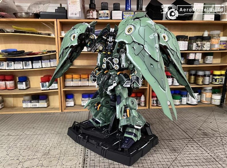May include: A detailed, green and black model robot with large wing-like structures. The model is on a black base, showcasing intricate mechanical details. The background includes shelves with various paints and model-building supplies.