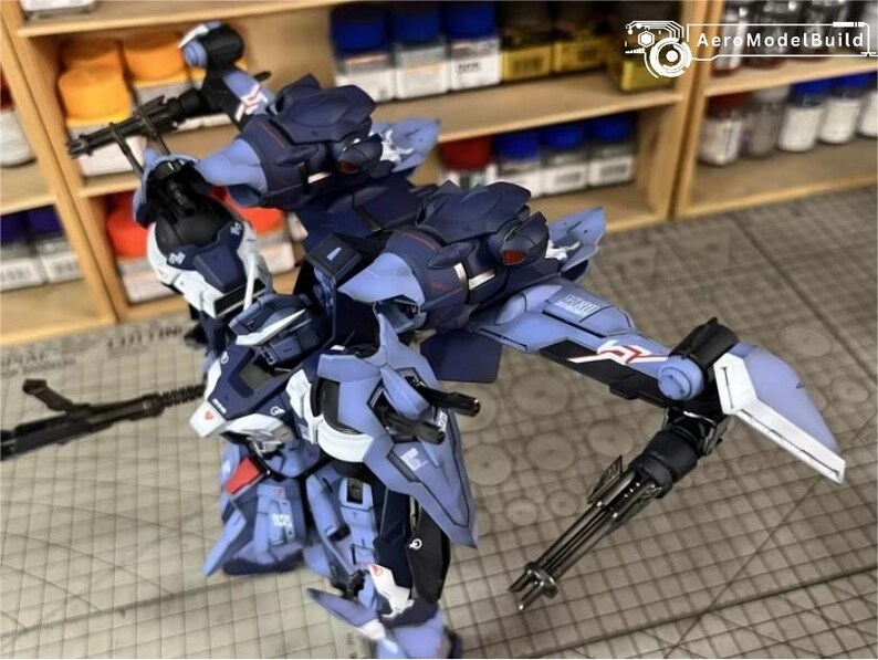 May include: A detailed, partially assembled model kit of a futuristic robot, primarily in shades of blue, white, and black. The model features intricate mechanical details, including articulated limbs and weapon attachments. The background shows shelves with paint bottles.