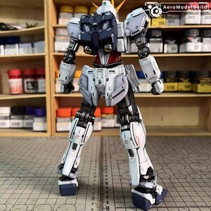 May include: A detailed, assembled model robot with a white, navy, and red color scheme. The robot has articulated joints and intricate panel lines, with a detailed head and shoulders. The model is posed on a grid surface, with paint bottles visible in the background.