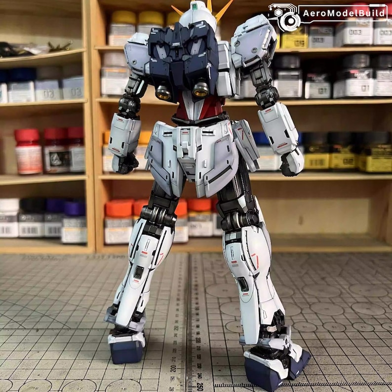 May include: A detailed, assembled model robot with a white, navy, and red color scheme. The robot has articulated joints and intricate panel lines, with a detailed head and shoulders. The model is posed on a grid surface, with paint bottles visible in the background.