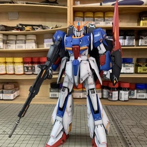May include: A detailed model robot in white, blue, red, and black. It holds a black rifle and a red blade. The model stands on a grid surface, with shelves and paint bottles behind it. The model is a detailed representation of a robot.