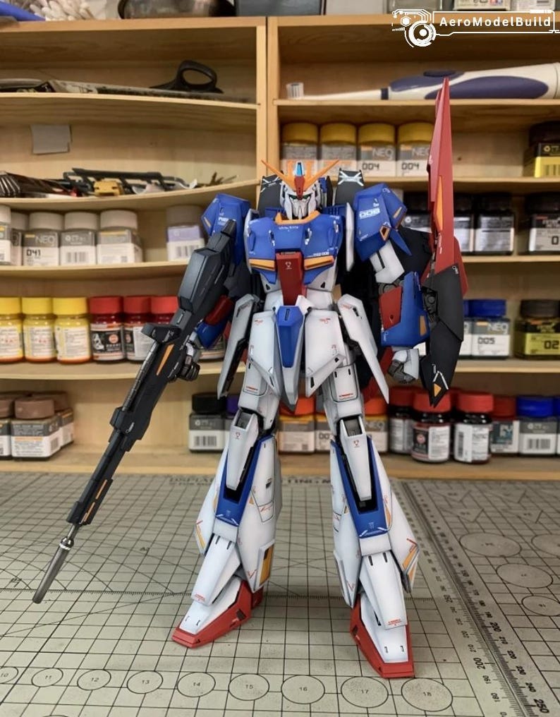 May include: A detailed model robot in white, blue, red, and black. It holds a black rifle and a red blade. The model stands on a grid surface, with shelves and paint bottles behind it. The model is a detailed representation of a robot.
