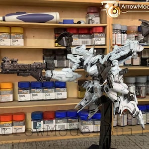 Könnte beinhalten: Detailliertes Modell eines weißen und schwarzen Mecha-Roboters, ausgestellt auf einem schwarzen Ständer. Das Modell befindet sich vor einem Holzregal mit Farbtöpfen und Werkzeugen. Der Text "ArrowModelBuild" ist sichtbar.