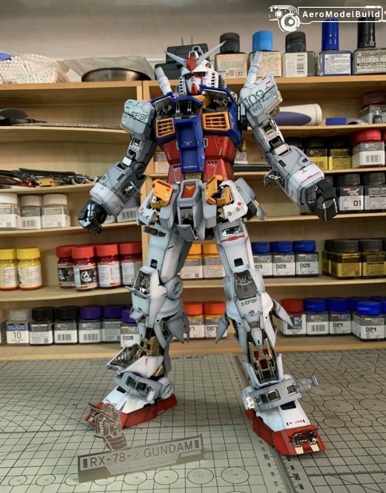 K&ouml;nnte beinhalten: Ein detailliertes Modell des RX-78-2 Gundam, ein Mecha-Roboter, wird gezeigt. Das Modell hat ein Farbschema aus Wei&szlig;, Blau, Rot und Gelb. Das Modell steht auf einer karierten Oberfl&auml;che, mit Farbt&ouml;pfen und Regalen im Hintergrund. Der Text "RX-78-2 GUNDAM" ist sichtbar.
