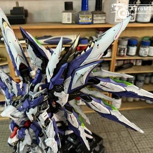 May include: A detailed model of a futuristic robot with large, stylized wings. The model features a colour scheme of white, blue, and purple with green accents. The model is on a black base, with the AeroModelBuild logo visible in the background.