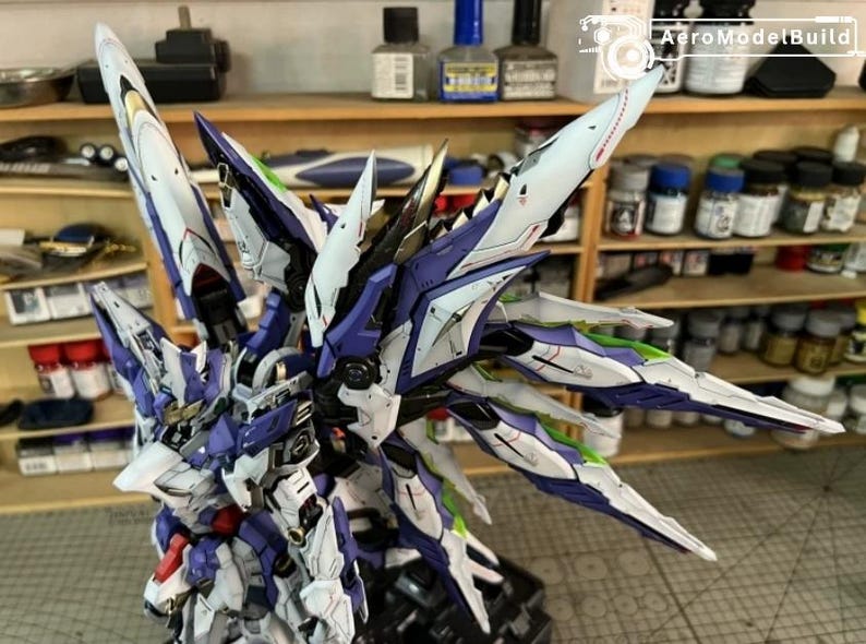 May include: A detailed model of a futuristic robot with large, stylized wings. The model features a colour scheme of white, blue, and purple with green accents. The model is on a black base, with the AeroModelBuild logo visible in the background.