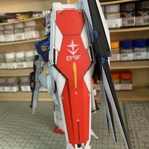 K&ouml;nnte beinhalten: Detailliertes Modell eines Mecha-Roboters, haupts&auml;chlich wei&szlig;, rot und blau. Der Roboter hat einen gro&szlig;en Schild mit dem Text "EFSF" und einem Sternemblem. Das Modell steht auf einer Rasteroberfl&auml;che, im Hintergrund Regale mit Farbe und Zubeh&ouml;r.