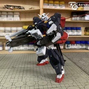 Puede incluir: Modelo detallado de un robot mecha, con cabeza blanca, detalles rojos y armadura azul oscuro. El robot sostiene un arma grande y est&aacute; sobre una superficie cuadriculada. El fondo incluye estantes con botes de pintura.