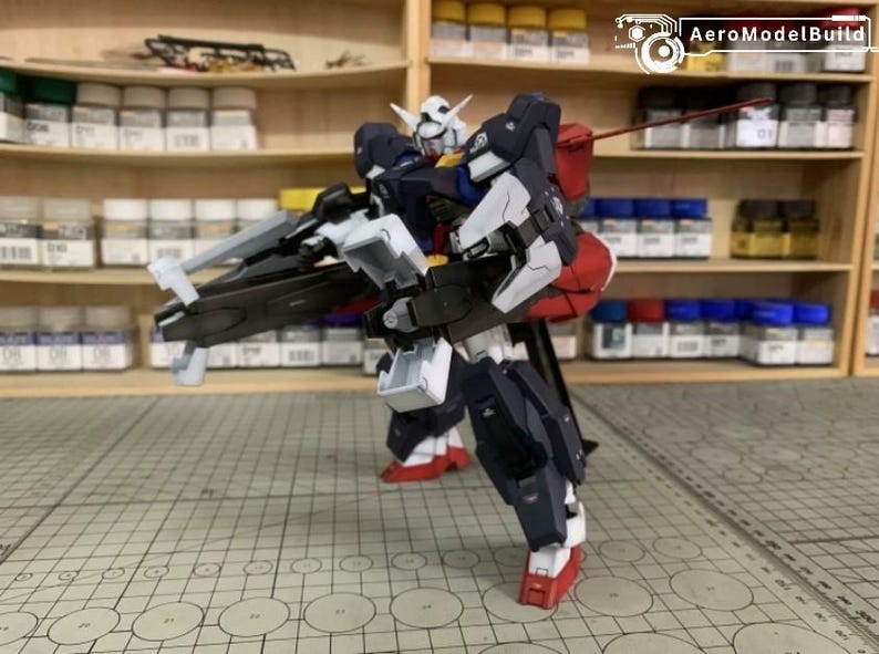 Puede incluir: Modelo detallado de un robot mecha, con cabeza blanca, detalles rojos y armadura azul oscuro. El robot sostiene un arma grande y est&aacute; sobre una superficie cuadriculada. El fondo incluye estantes con botes de pintura.