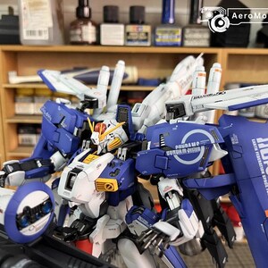 May include: A detailed model of a Gundam robot, predominantly blue, white, and yellow. The model features intricate designs, including wings and a shield with text. The robot is set against a background of shelves with paint supplies.