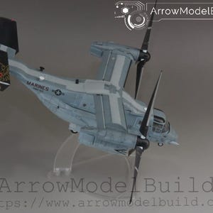 May include: A detailed model of a military aircraft, featuring a gray and white color scheme with black propellers. The model is labeled "Marines" and includes the text "ArrowModelBuild". The aircraft is mounted on a clear stand.