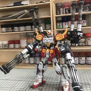 K&ouml;nnte beinhalten: Detailliertes Modell eines Gundam-Roboters mit wei&szlig;em, rotem, gelbem und schwarzem Farbschema. Der Roboter ist mit mehreren gro&szlig;en Kanonen ausgestattet und befindet sich in einer dynamischen Pose. Der Hintergrund zeigt Regale mit Farbt&ouml;pfen und Modellbauzubeh&ouml;r.