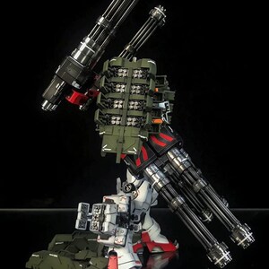 K&ouml;nnte beinhalten: Eine wei&szlig;e, rote und schwarze Roboter-Actionfigur mit zwei gro&szlig;en mehrl&auml;ufigen Gatling-Kanonen, einem gr&uuml;nen Raketenwerfer-Rucksack und an den Beinen montierten Waffensystemen. Der Text AeroModelBuild ist oben rechts zu sehen.