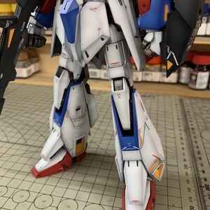 May include: A detailed model robot with white, blue, red, and black armour. The robot has intricate panel lines and markings, with a matte finish. The image includes the text "AeroModelBuild".