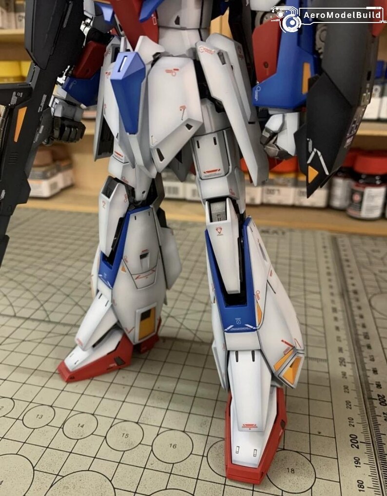 May include: A detailed model robot with white, blue, red, and black armour. The robot has intricate panel lines and markings, with a matte finish. The image includes the text "AeroModelBuild".