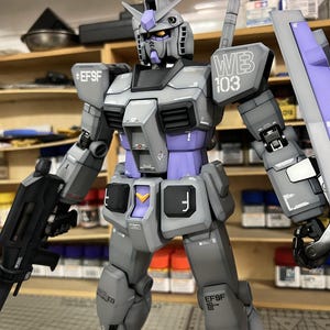 May include: A detailed model of a Gundam robot, primarily gray with purple accents. The robot is holding a black weapon and a shield. The text "WEB 103" is visible on the shoulder. The model is a collectible item.