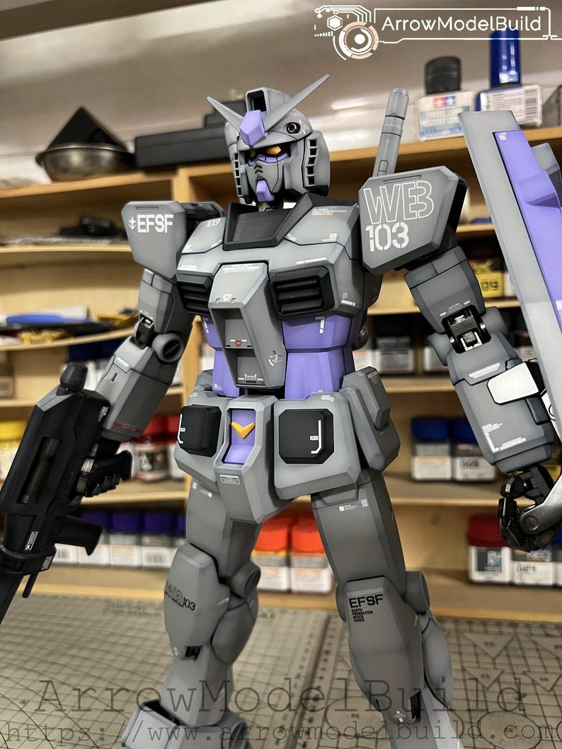 May include: A detailed model of a Gundam robot, primarily gray with purple accents. The robot is holding a black weapon and a shield. The text "WEB 103" is visible on the shoulder. The model is a collectible item.