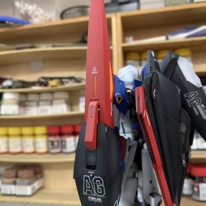 May include: A partially assembled model kit with a red and black shield, displaying the letters "AG" and the text "AEUG". The model has blue, white, and yellow details. Shelves with bottles and containers are visible in the background.