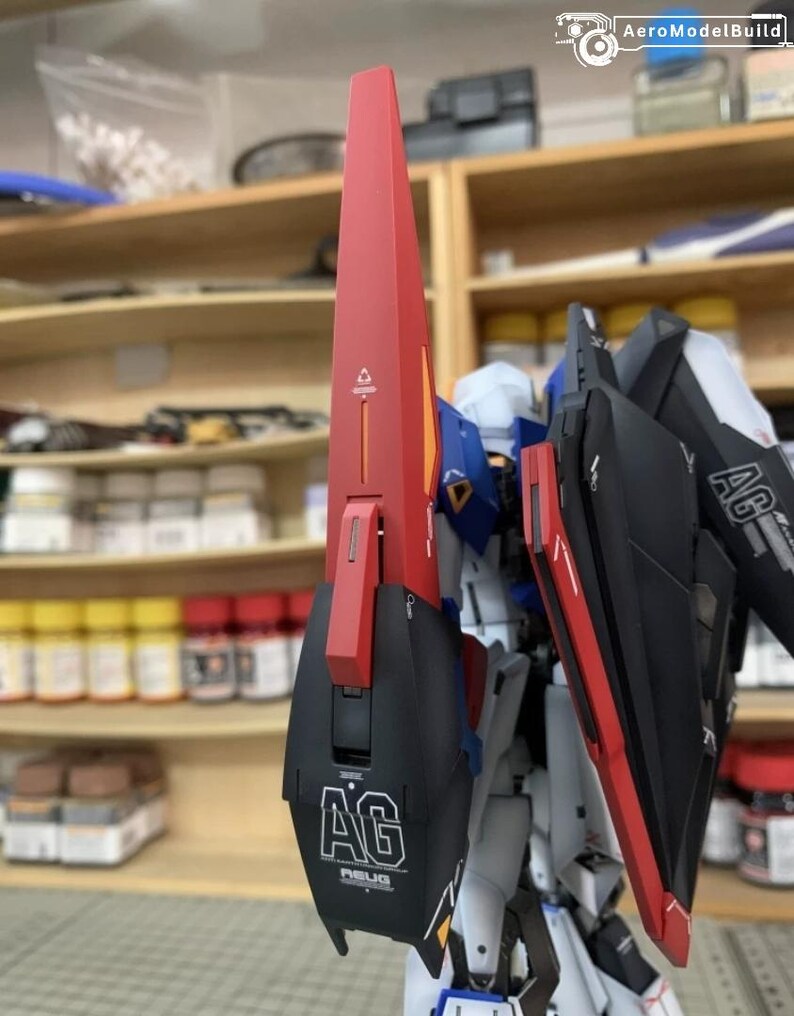 May include: A partially assembled model kit with a red and black shield, displaying the letters "AG" and the text "AEUG". The model has blue, white, and yellow details. Shelves with bottles and containers are visible in the background.