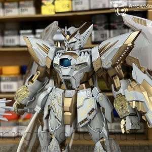 May include: A detailed model of a white and gold mecha robot with intricate designs. The robot has large wings, a blue orb on its chest, and is holding a weapon. The model is set against a blurred background of shelves.