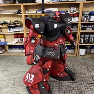 May include: A detailed model of a red and black robot with the text "A12" and "PRINCIPALITY OF ZEON". The robot has multiple articulated parts and is posed on a grid surface. The model is set against a backdrop of shelves filled with paint bottles.