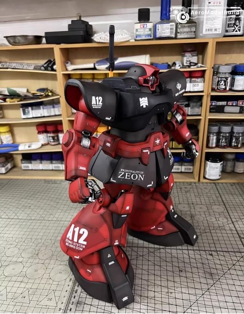 May include: A detailed model of a red and black robot with the text "A12" and "PRINCIPALITY OF ZEON". The robot has multiple articulated parts and is posed on a grid surface. The model is set against a backdrop of shelves filled with paint bottles.