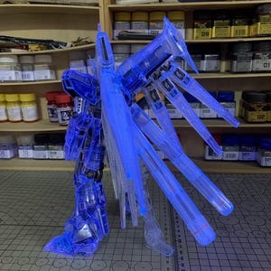 May include: A transparent blue model robot kit, partially assembled, with large wing-like structures. The model is lit with blue light and stands on a grid-patterned surface. The kit is likely for display or hobby purposes.