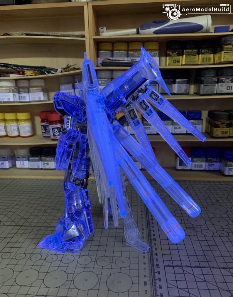 May include: A transparent blue model robot kit, partially assembled, with large wing-like structures. The model is lit with blue light and stands on a grid-patterned surface. The kit is likely for display or hobby purposes.