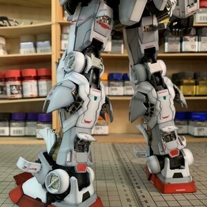K&ouml;nnte beinhalten: Detailliertes, teilweise zusammengebautes Modell eines Roboters mit wei&szlig;en, grauen und roten Akzenten. Die Beine und der untere Torso des Roboters sind sichtbar und zeigen komplizierte mechanische Details. Das Modell steht auf einer Rasteroberfl&auml;che, mit Farbt&ouml;pfen im Hintergrund.