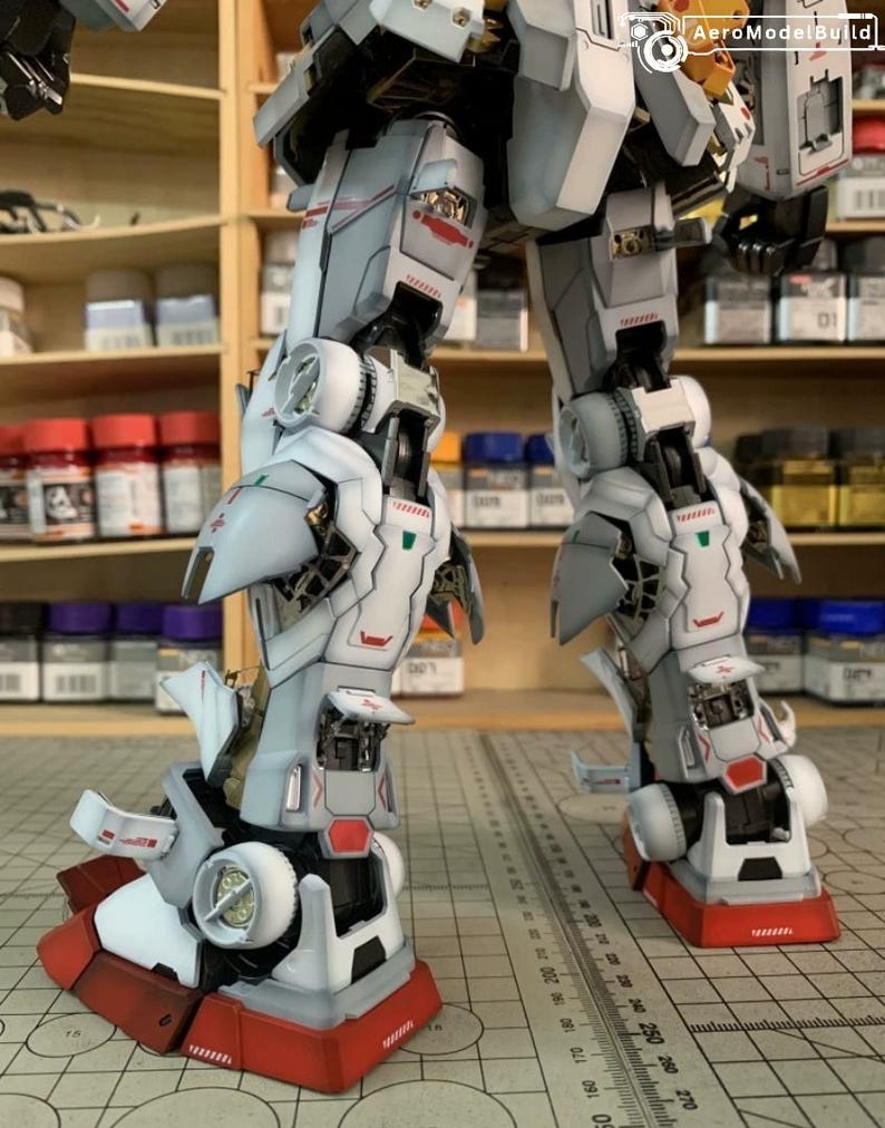 K&ouml;nnte beinhalten: Detailliertes, teilweise zusammengebautes Modell eines Roboters mit wei&szlig;en, grauen und roten Akzenten. Die Beine und der untere Torso des Roboters sind sichtbar und zeigen komplizierte mechanische Details. Das Modell steht auf einer Rasteroberfl&auml;che, mit Farbt&ouml;pfen im Hintergrund.