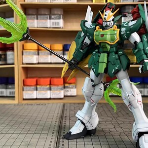 May include: A detailed model of a Gundam robot, predominantly green and white, with yellow and red details. It holds a green energy weapon. The model is posed on a grid surface, with paint bottles and shelves in the background. The text "ArrowModelBuild" is visible.