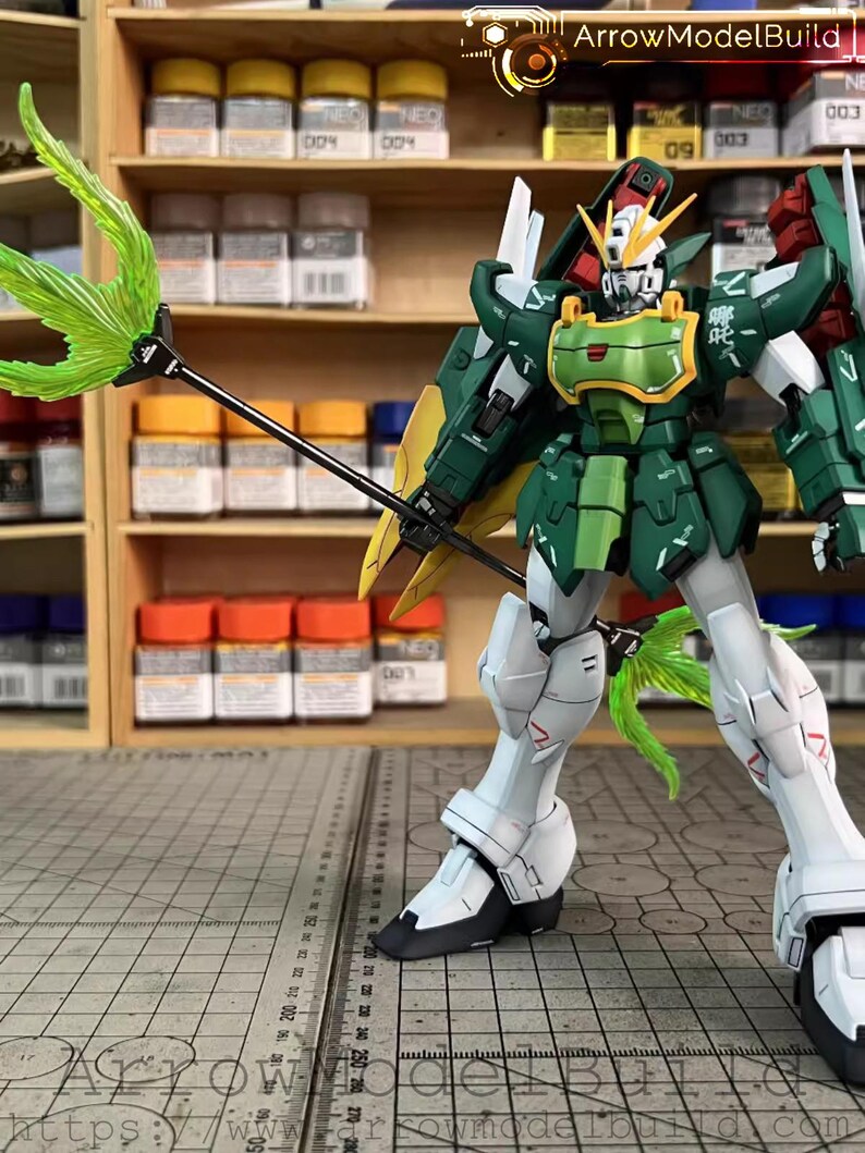May include: A detailed model of a Gundam robot, predominantly green and white, with yellow and red details. It holds a green energy weapon. The model is posed on a grid surface, with paint bottles and shelves in the background. The text "ArrowModelBuild" is visible.