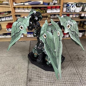 May include: A detailed model of a mechanical figure with large, stylized wings. The model is primarily green and black, with intricate paneling and circular accents. It is displayed on a black base, suggesting a collectible or display piece.