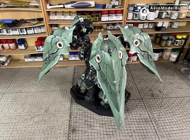 May include: A detailed model of a mechanical figure with large, stylized wings. The model is primarily green and black, with intricate paneling and circular accents. It is displayed on a black base, suggesting a collectible or display piece.