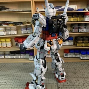 K&ouml;nnte beinhalten: Ein detailliertes Modell eines Gundam-Roboters, haupts&auml;chlich wei&szlig;, grau, rot und blau. Das Modell weist komplizierte mechanische Details auf, darunter bewegliche Gliedma&szlig;en und ein Rucksack mit zwei zylindrischen Komponenten. Der Roboter steht auf einer gemusterten Oberfl&auml;che, mit Regalen voller Farbe und Werkzeuge im Hintergrund.