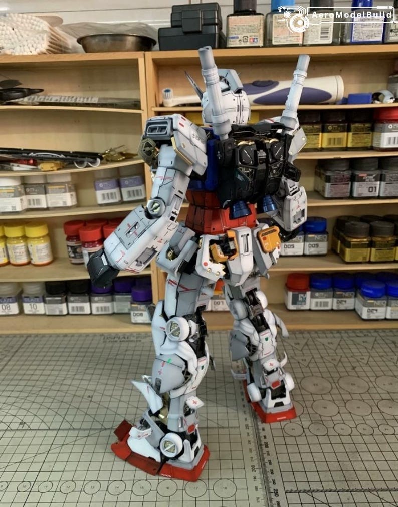 K&ouml;nnte beinhalten: Ein detailliertes Modell eines Gundam-Roboters, haupts&auml;chlich wei&szlig;, grau, rot und blau. Das Modell weist komplizierte mechanische Details auf, darunter bewegliche Gliedma&szlig;en und ein Rucksack mit zwei zylindrischen Komponenten. Der Roboter steht auf einer gemusterten Oberfl&auml;che, mit Regalen voller Farbe und Werkzeuge im Hintergrund.