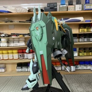 Puede incluir: Kit de modelo AeroModelBuild de un robot mecha verde y rojo con un gran escudo. El modelo est&aacute; parcialmente ensamblado y presenta detalles intrincados, incluido un emblema triangular en el escudo y un soporte negro. Fondo con estantes con botes de pintura.