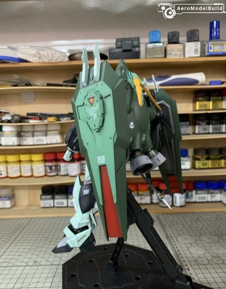 Puede incluir: Kit de modelo AeroModelBuild de un robot mecha verde y rojo con un gran escudo. El modelo est&aacute; parcialmente ensamblado y presenta detalles intrincados, incluido un emblema triangular en el escudo y un soporte negro. Fondo con estantes con botes de pintura.