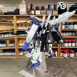 May include: A detailed model kit of a mecha robot, mainly white, blue, and black. The model is displayed on a clear stand, highlighting its intricate design. The background shows shelves filled with paint bottles and tools, indicating a hobbyist's workspace.