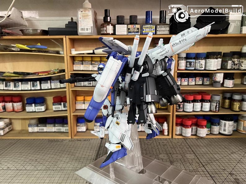 May include: A detailed model kit of a mecha robot, mainly white, blue, and black. The model is displayed on a clear stand, highlighting its intricate design. The background shows shelves filled with paint bottles and tools, indicating a hobbyist's workspace.