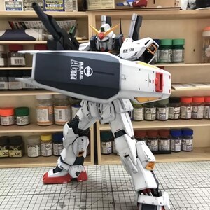 Op de afbeelding: Een gedetailleerd model van een witte, blauwe en rode robot met de tekst "AEUG AG004" op zijn schild. De robot poseert met &eacute;&eacute;n been uitgestrekt, waarbij ingewikkelde mechanische details worden getoond. Het model staat tegen een achtergrond van verfbenodigdheden.